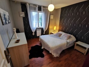 A bedroom with a bed, bedside tables, a desk, a window with curtains, a radiator, a hanging light fixture, and a patterned wall. - Modern Cottage near Blois Chambord with Private Terrace, Garden, and Wi-Fi (Les Montils)