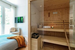 Een kamer met een bed, een houten sauna, een glazen scheidingswand en uitzicht op groen.