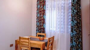 Een eetruimte met een ronde houten tafel, stoelen, een geweven vloerkleed, een hanglamp en lichte gordijnen met gestreept gordijn.