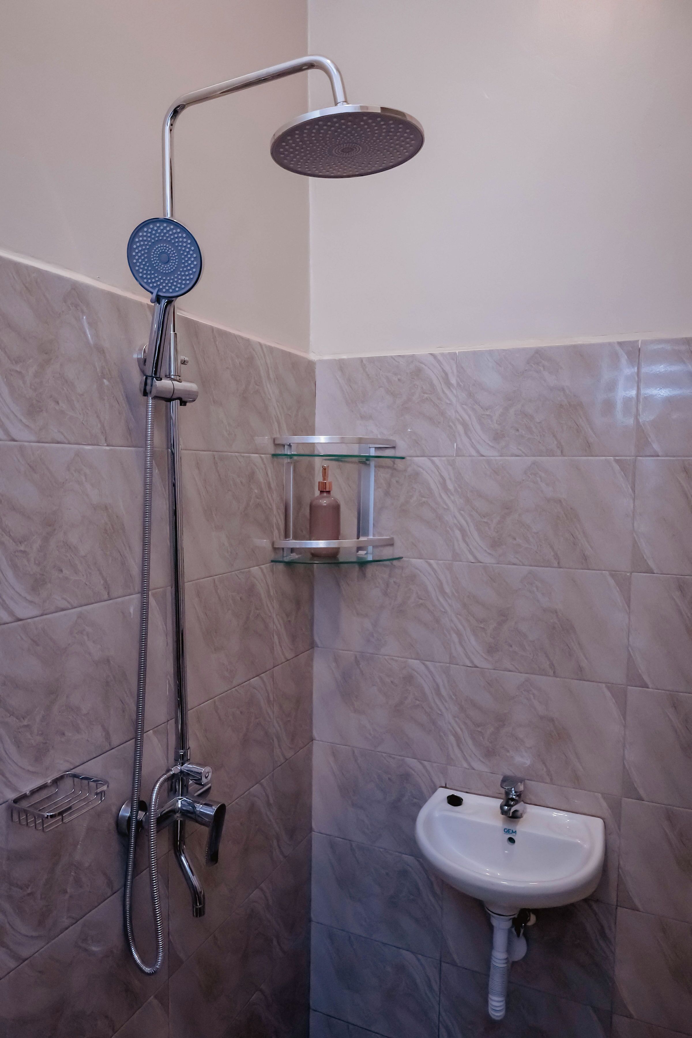 Een badkamer met een douche, wastafel en een glazen plankje met een fles.