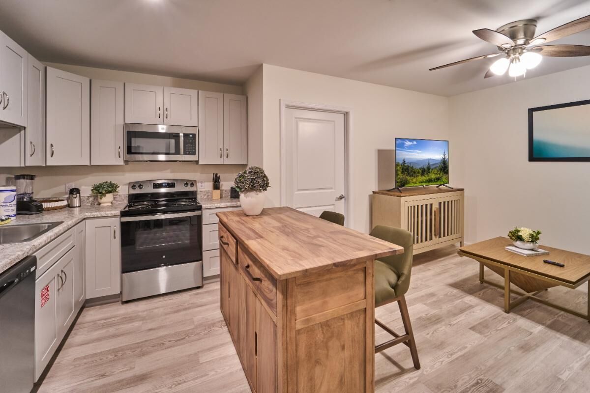 A modern kitchen with white cabinets, stainless steel appliances, a wooden island, and a dining area with a TV and plant.
