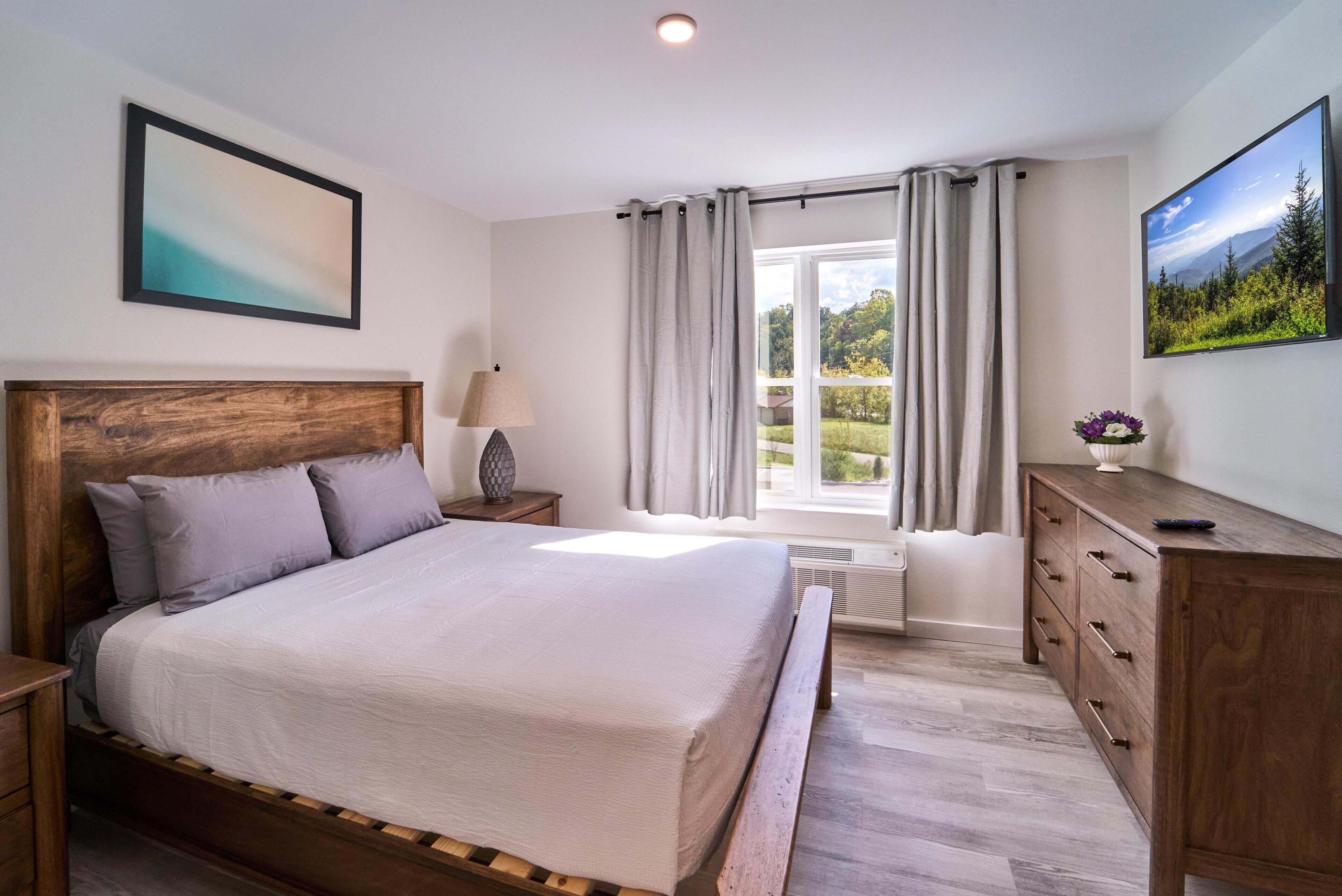 A bedroom with a wooden bed, a nightstand, a dresser, a window with curtains, and a painting on the wall.