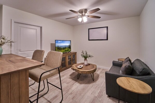 A modern living room with a wooden dining table, two chairs, a sofa, a coffee table, a television, and a ceiling fan. - The Ruby 205 (Pigeon Forge)