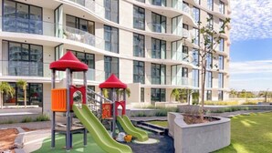 A playground with slides and climbing structures in front of a modern building with multiple floors and balconies.