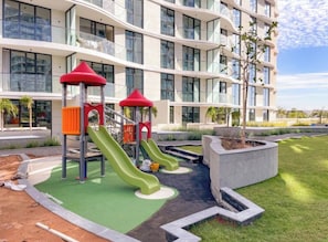 Un parque infantil con toboganes y estructuras para escalar frente a un edificio moderno de varios pisos y balcones.