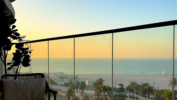 A balcony with a view of the beach and ocean, a chair, a table, and a plant.