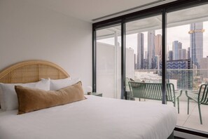 A hotel room with a large bed, a view of a cityscape through floor-to-ceiling windows, and a balcony with chairs. - 2BD | Immaculate SkyHigh Apartment |Prime Location (Southbank)