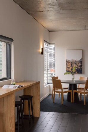 A modern interior with a wooden desk, black stools, a round table with chairs, a cityscape painting, and a window with blinds. - 2BD | Immaculate SkyHigh Apartment |Prime Location (Southbank)