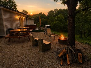 Eine Terrasse mit Feuerstelle, Picknicktisch und Stühlen, umgeben von Bäumen und einem Gebäude.