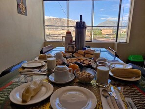 Een ontbijttafel met borden, melk, gebak, een koffiezetapparaat en een uitzicht op bergen door een raam.