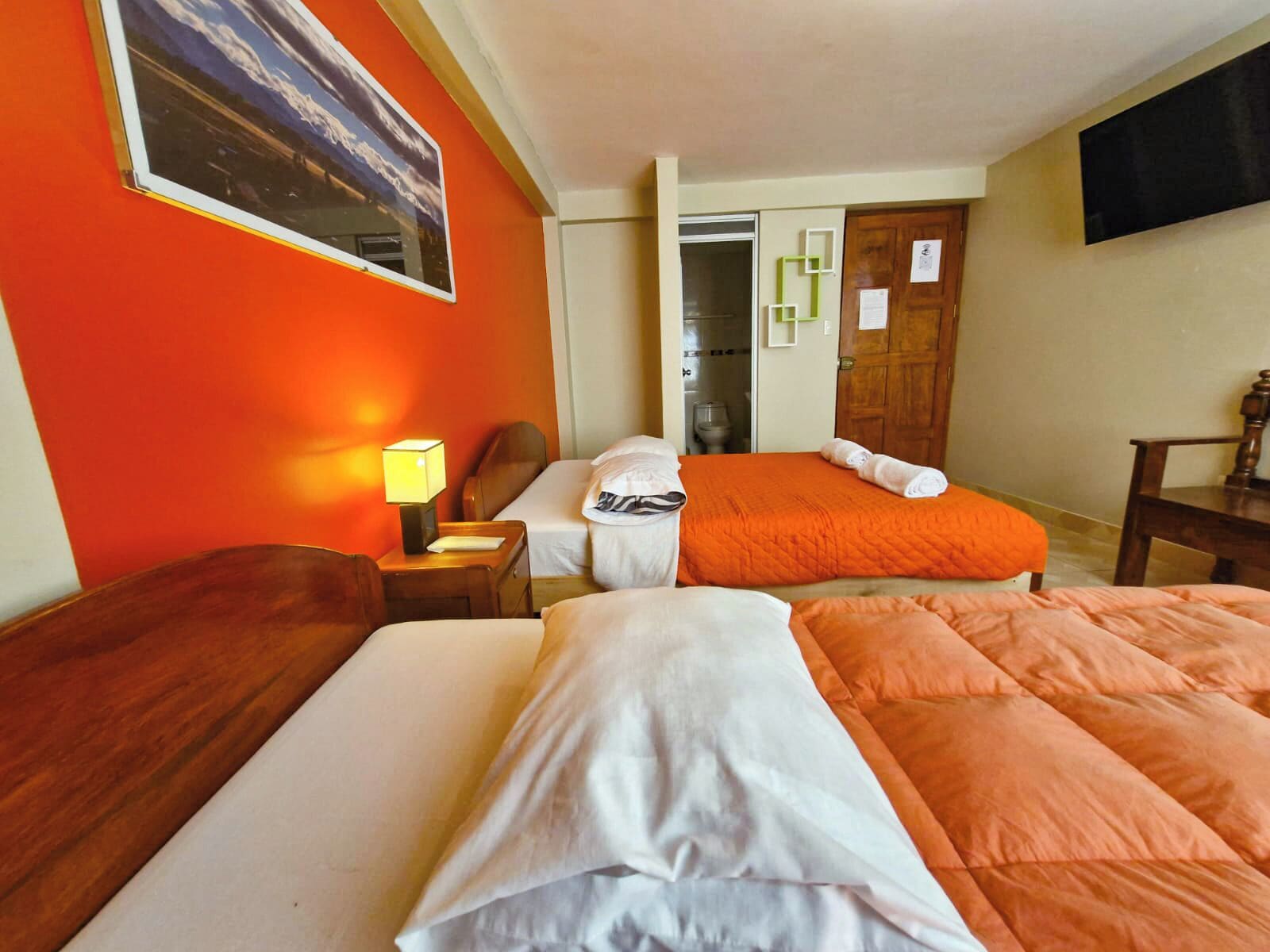 Une chambre d’hôtel avec deux lits, une table de chevet, une lampe, une chaise en bois, un téléviseur fixé au mur et une porte avec un miroir.