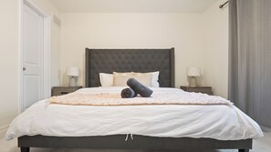 Un dormitorio con una cama grande, dos mesitas de noche con lámparas, un cabecero oscuro y una puerta.