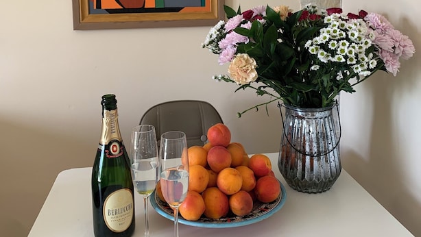 Une table avec une bouteille de champagne, des verres, une assiette de pêches et un vase de fleurs.
