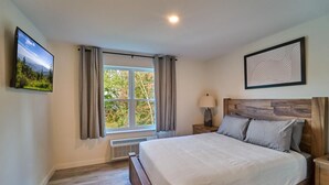 Een slaapkamer met een groot bed, een raam met gordijnen, een wandtelevisie, een bedlampje en een ingelijste foto.