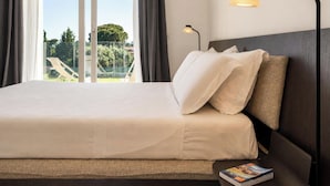 Habitación de hotel con cama, mesita de noche, lámpara, ventana con cortinas y vistas a zonas verdes.