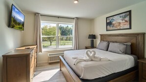 Ein Schlafzimmer mit einem Bett, einem Fernseher, einem Fenster mit Vorhängen, einem Bild und einem Nachttisch.