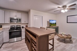 Eine Küche mit Mikrowelle, Herd und Weinkühlschrank, ein Essbereich mit Tisch und Stühlen, ein Deckenventilator und ein Fernseher auf einem Schrank.