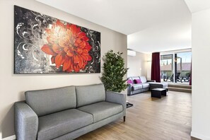 A living room with a grey sofa, a large floral painting, a green plant, and a view through a glass door. - Lake Garda Nidesti Suite Apartment (DESENZANO DEL GARDA)