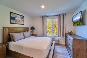 Um quarto com cama, mesas de cabeceira, cômoda, janela com cortinas e televisão presa na parede.