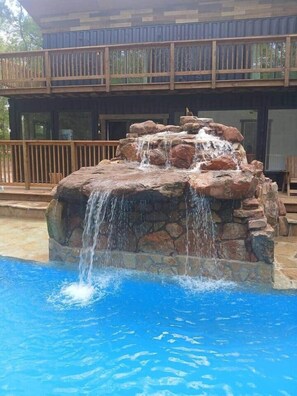 Piscina com cascata, deck de madeira e um edifício com varanda ao fundo.
