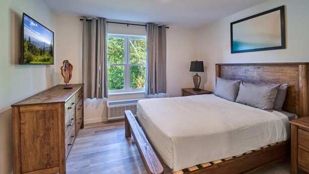 Ein Schlafzimmer mit einem Bett, einem Nachttisch aus Holz, einem Beistelltisch, einem Fernseher, einem Fenster mit Vorhängen und einem gerahmten Bild an der Wand.