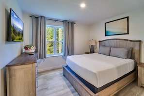 Un dormitorio con cama, mesita de noche, lámpara, televisor, ventana con cortinas e imagen en la pared.