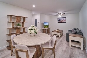Ein modernes Wohnzimmer mit einem runden Holztisch, beigen Sesseln, einem Fernseher, einer Couch und einem Deckenventilator.