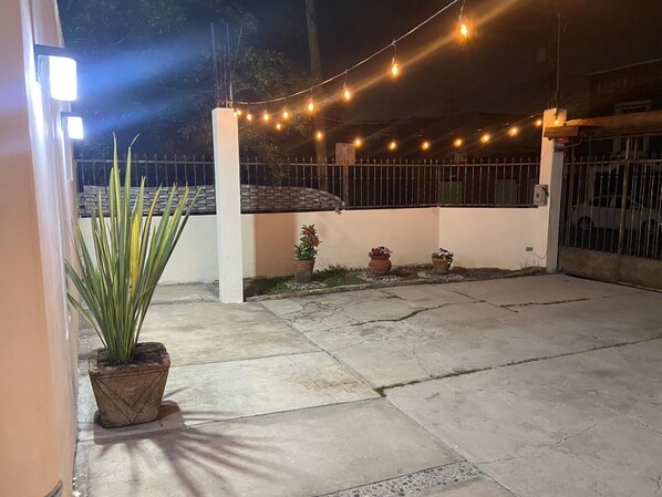 An outdoor area with a concrete floor, potted plants, a metal gate, and string lights. - Full House Steps from BBVA Stadium (Guadalupe)