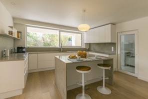 A modern kitchen with a large window, a central island with stools, and a built-in refrigerator. - Golf Escape Villa by Cadenzalux (Sintra)