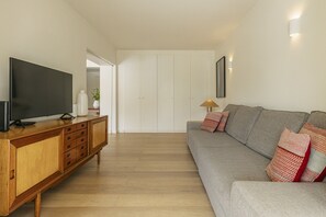 Una sala de estar moderna con un televisor de pantalla plana sobre un mueble de madera, un sofá gris con cojines estampados y un armario blanco al fondo.