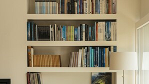 Ein Bücherregal mit Büchern, ein Holzstuhl, eine Vase mit getrockneten Blumen und eine Lampe.