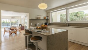 Eine moderne Küche mit einer zentralen Kochinsel, Barhocker, einem Essbereich mit Holztisch und Stühlen sowie einem großen Fenster, das für natürliches Licht sorgt.