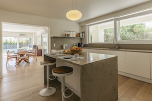 A modern kitchen with a central island, bar stools, a dining area with a wooden table and chairs, and a large window providing natural light. - Golf Escape Villa by Cadenzalux (Sintra)