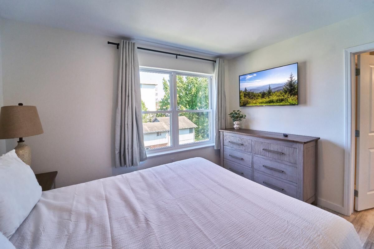 A bedroom with a large bed, a dresser, a television on the wall, and a window with curtains.