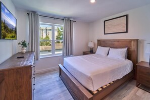 Ein Schlafzimmer mit Bett, Nachttischen, einem Schreibtisch, einem Fenster mit Vorhängen und Blick auf Gebäude.