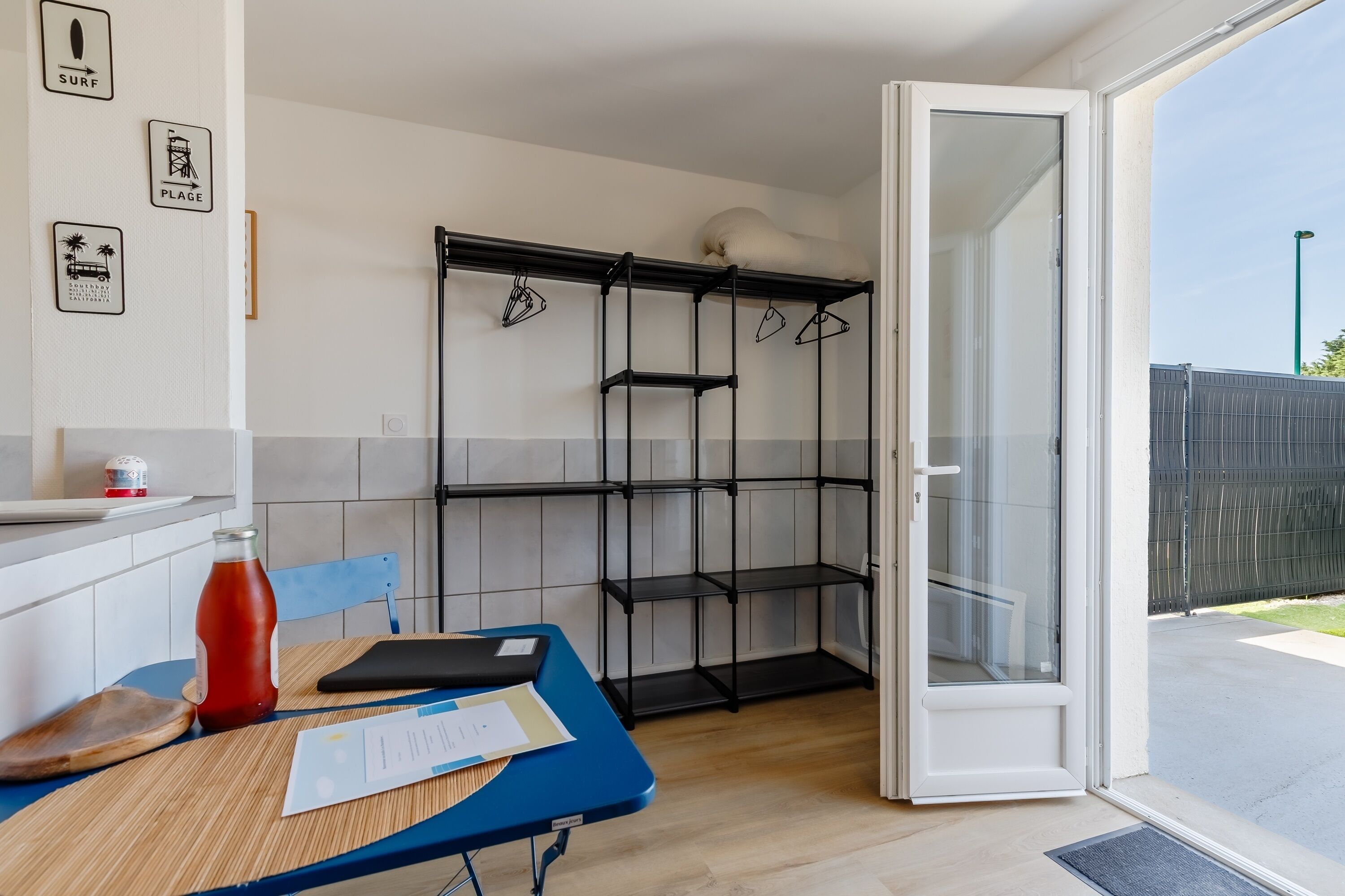 A kitchen with a blue table, a red bottle, and a black folder. There are shelves with hangers and a door leading outside.