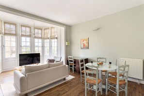 A living room with a sofa, TV, dining table, chairs, and large windows. - Les Ondines – Élégante maison de caractère au cœur de Saint-Lunaire (Saint-Lunaire)