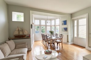 Une pièce comprenant une table à manger, des chaises, un canapé, un radiateur et une fenêtre donnant sur un espace verdoyant.