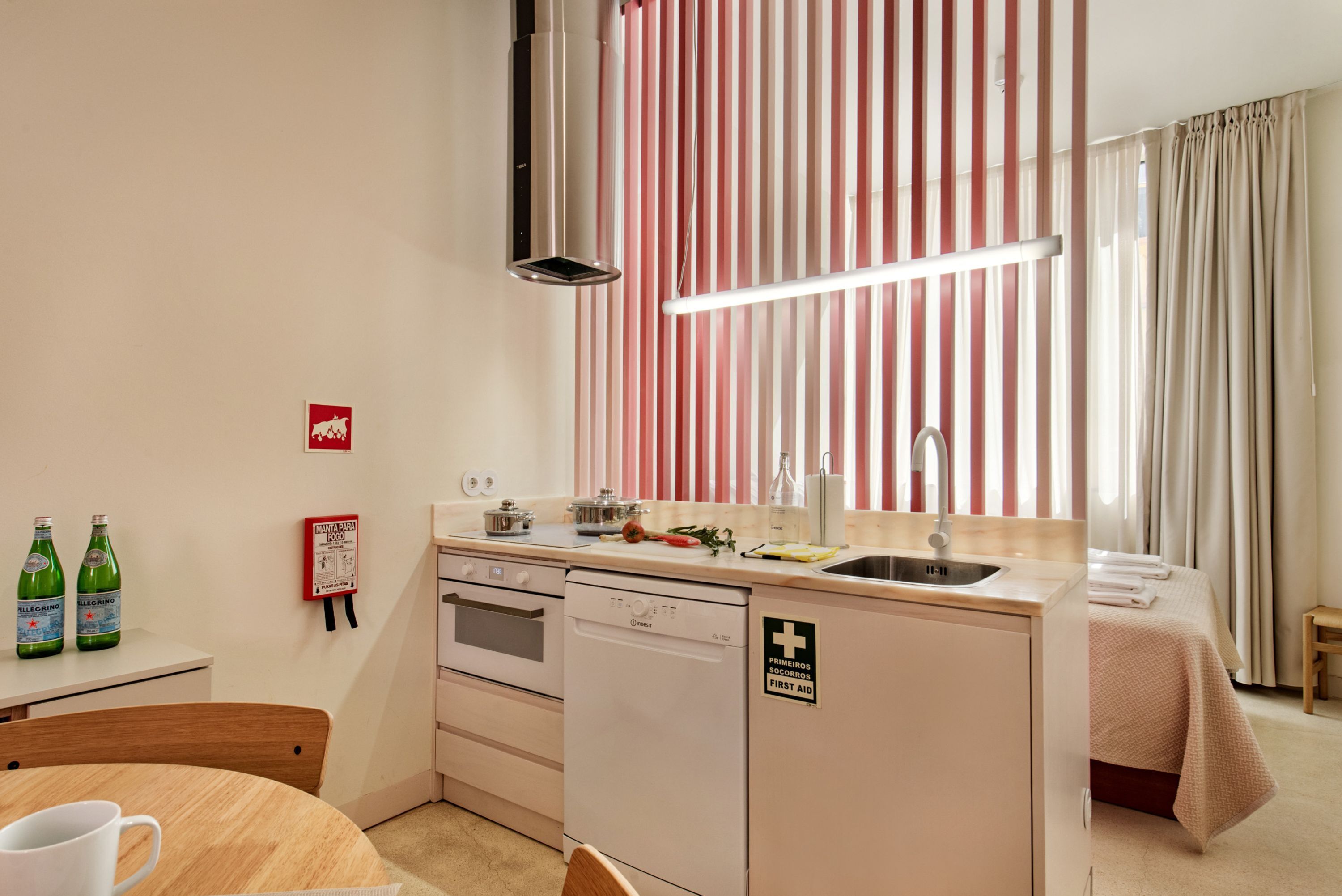 Una cocina compacta con lavavajillas, estufa y pileta. Hay una mesa pequeña con una taza y una botella de agua. Un detector de incendios está colocado en la pared.