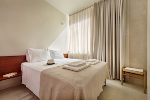 Una cama bien tendida con una colcha blanca y almohadas, una mesita de noche con un libro y un sombrero, y una ventana con cortinas.
