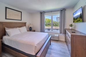 Un dormitorio con cama, mesita de noche, cómoda, ventana con cortinas y televisor instalado en la pared.