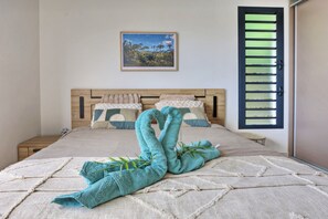 Ein ordentlich bezogenes Bett mit dekorativer Handtuchgestaltung, ein gerahmtes Bild an der Wand und ein Fenster mit Jalousien.