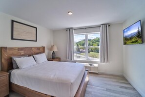A bedroom with a wooden headboard, a bed with white bedding, a nightstand with a lamp, a window with curtains, and a framed picture on the wall. - The Ruby 304 (Pigeon Forge)