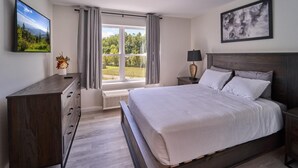 Une chambre à coucher comprenant un lit, une commode, une table de chevet, une lampe, une fenêtre avec des rideaux et un tableau accroché au mur.