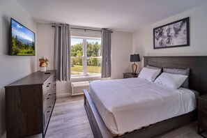 A bedroom with a bed, a dresser, a nightstand, a lamp, a window with curtains, and a painting on the wall. - The Ruby 306 (Pigeon Forge)