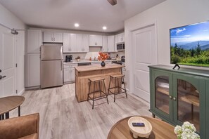 A modern kitchen with white cabinets, a wooden island with stools, a green cabinet with glass doors, and a TV displaying a landscape. - The Ruby 309 (Pigeon Forge)