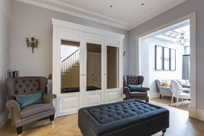 Ein geräumiges Wohnzimmer mit einem großen Pouf, zwei Sesseln, einem weißen Schrank mit Türen und einer Treppe, die zu einer weiteren Ebene führt.