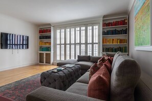 Uma sala de estar com um sofá cinza, um tapete vermelho e preto, uma estante e um quadro na parede.