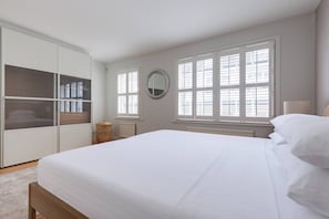 Ein Schlafzimmer mit einem großen Bett, einem Kleiderschrank, zwei Fenstern mit Jalousien, einem runden Spiegel und einer Nachttischlampe.