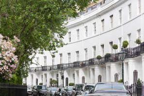 Ein weißes Gebäude mit zahlreichen Fenstern und Balkonen, davor parkende Autos und ein Baum mit rosa Blüten.